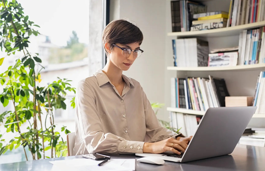 Woman working on a laptop in a home office setting with plants and books in the background.